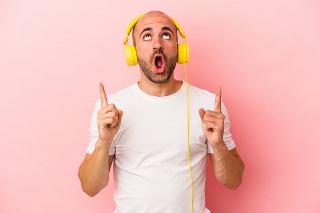 Jeune homme chauve caucasien écoutant de la musique isolée sur fond rose pointant vers le haut avec la bouche ouverte.