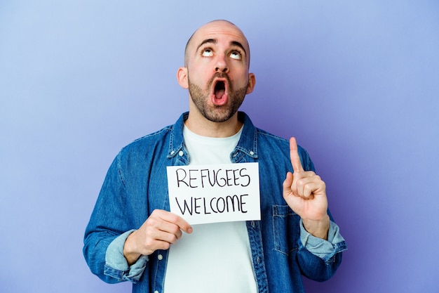Jeune homme chauve caucasien tenant une pancarte de bienvenue de réfugiés isolée sur un mur bleu pointant vers le haut avec la bouche ouverte.