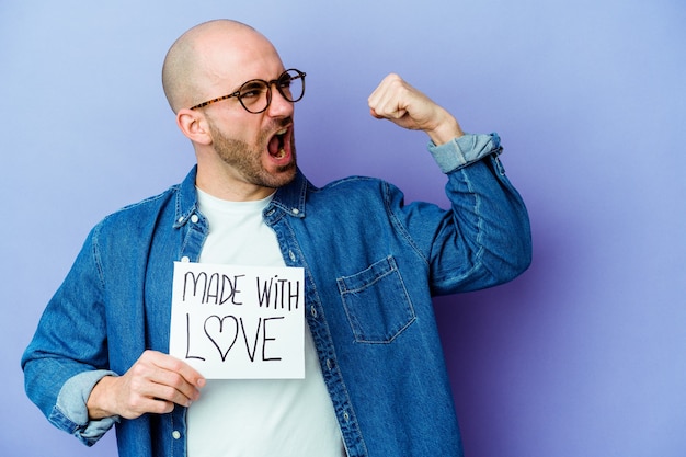 Photo jeune homme chauve caucasien tenant une pancarte faite avec amour isolé sur mur violet levant le poing après une victoire, concept gagnant.