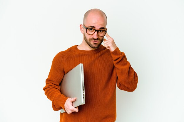 Jeune homme chauve tenant un ordinateur portable sur blanc montrant un geste de déception avec l'index.