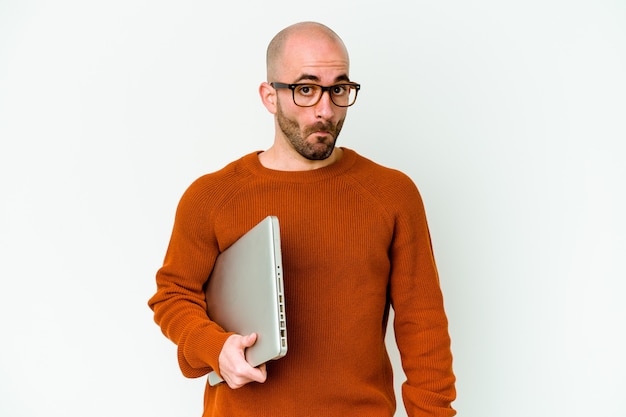 Jeune homme chauve tenant un ordinateur portable isolé sur fond blanc hausse les épaules et ouvre les yeux confus.