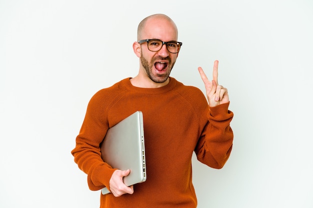 Jeune homme chauve tenant un ordinateur portable isolé sur un mur blanc joyeux et insouciant montrant un symbole de paix avec les doigts.