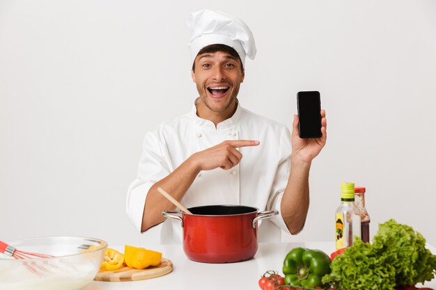 Jeune homme chef isolé sur un mur blanc faisant cuire à l'aide d'un téléphone portable pointant vers l'affichage.