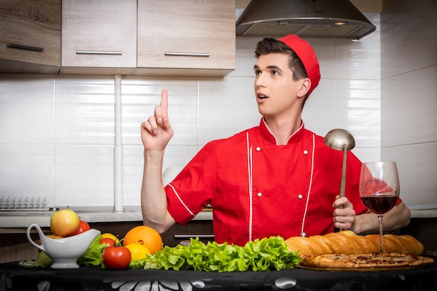 Jeune homme chef en uniforme rouge tenant l'index et la louche avec une excellente nouvelle idée sur fond clair