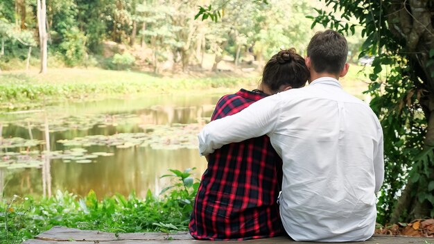 Jeune homme en chemise blanche, assis sur un banc près d'une femme aux cheveux noirs et s'embrasse avec amour contre le lac par une chaude journée ensoleillée en gros plan