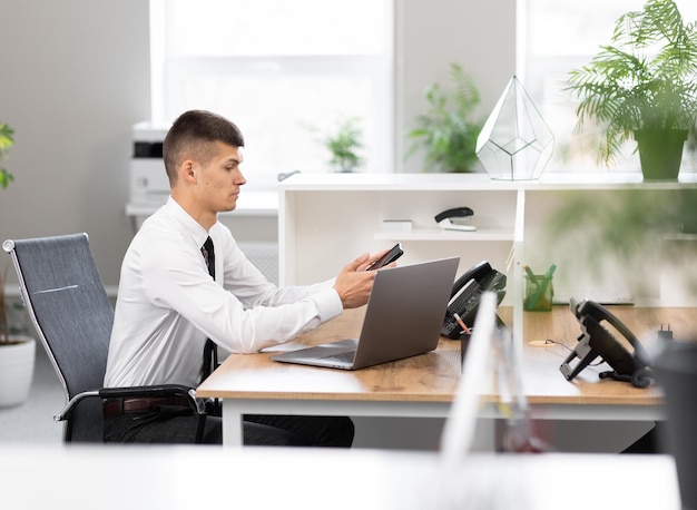 Jeune homme en chemise blanche avec une cravate noire au bureau au bureau lumineux travaillant sur un ordinateur portable