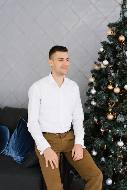 Un jeune homme en chemise blanche est assis sur un canapé près d'un arbre de Noël et sourit