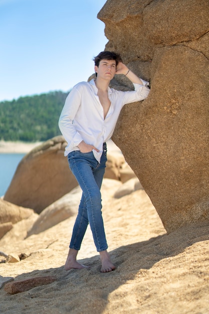 Jeune homme en chemise blanche et jeans appuyé contre un rocher