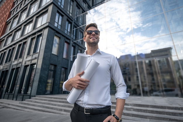 Jeune homme en chemise blanche et lunettes de soleil tenant des plans