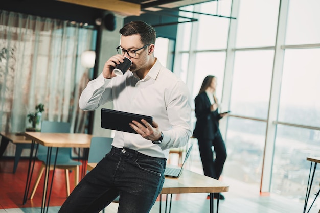 Un jeune homme en chemise blanche se tient dans un bureau moderne spacieux avec du café et travaille sur une tablette Un employé de bureau moderne Un homme d'affaires en lunettes travaille en ligne et boit du café chaud détendu