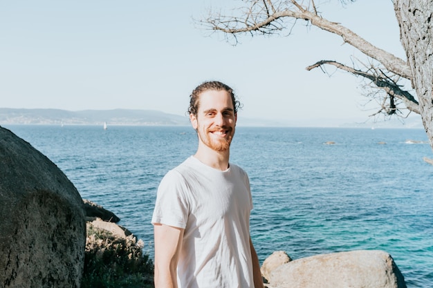 Jeune homme avec une chemise blanche vierge souriant à la caméra en toute confiance sur la côte espagnole lors d'une journée à la plage