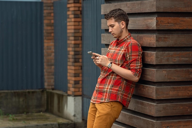 un jeune homme en chemise à carreaux se tient appuyé contre le mur et regarde le téléphone