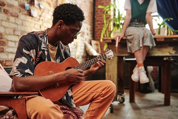 Jeune homme en chemise décontractée et pantalon jouant de la guitare assis dans un fauteuil