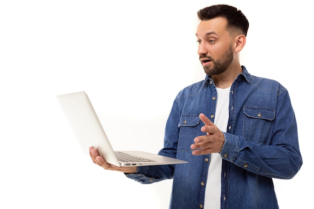 Un jeune homme en chemise en jean bleu tient un ordinateur portable dans ses mains et regarde l'écran avec surprise