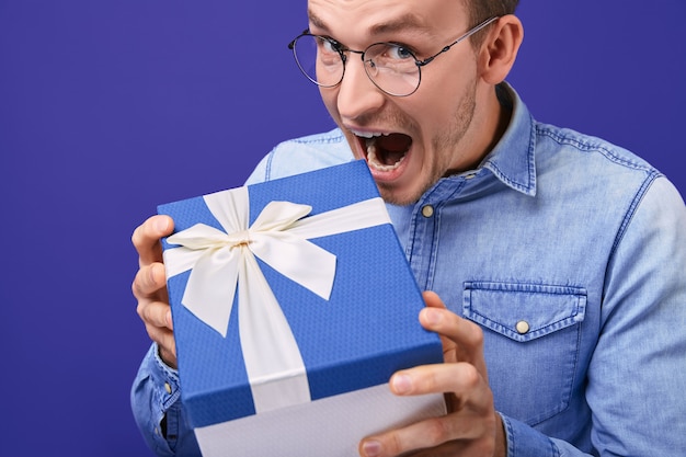 Jeune homme en chemise en jean et lunettes debout avec un cadeau avec un bonnet bleu dans ses mains