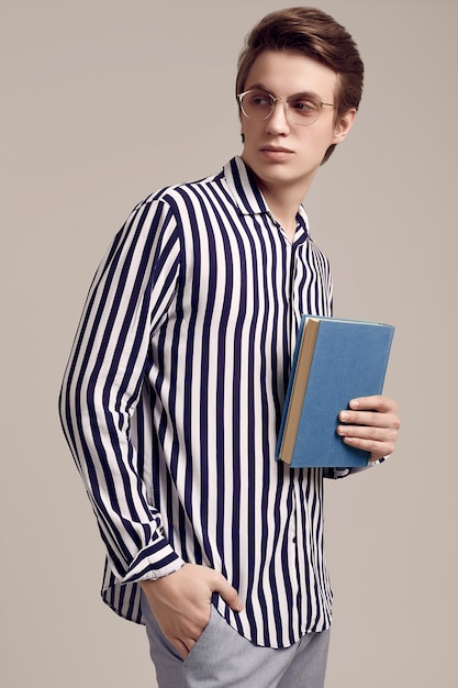 Jeune homme en chemise à rayures posant avec un livre sur fond gris