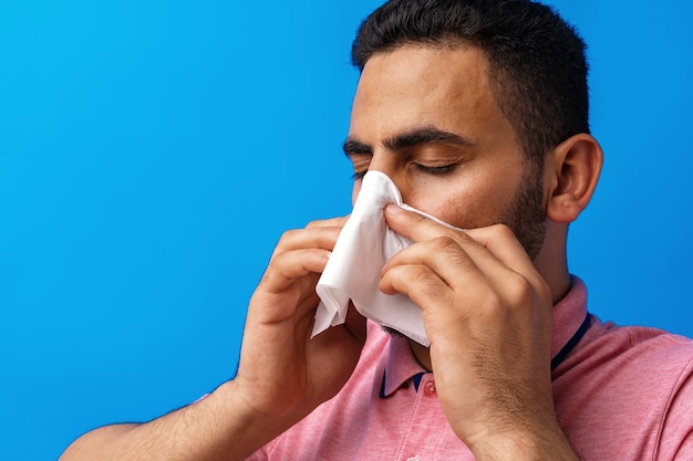 Jeune homme en chemise rose avec allergie ou froid se mouchant dans un mouchoir sur fond bleu