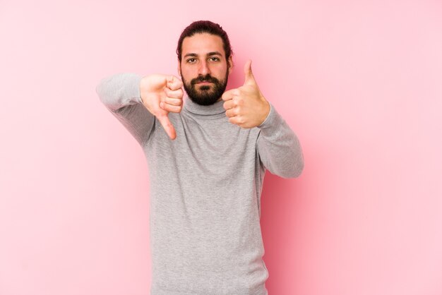 Jeune homme cheveux longs isolé sur un mur rose montrant les pouces vers le haut et les pouces vers le bas, difficile de choisir le concept