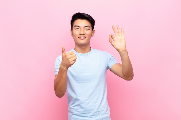 Jeune homme chinois se sentant heureux, étonné, satisfait et surpris, montrant les gestes corrects et les pouces vers le haut, souriant contre le mur de couleur plat