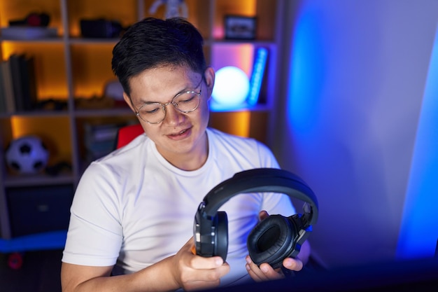 Jeune homme chinois streamer souriant confiant tenant des écouteurs dans la salle de jeux