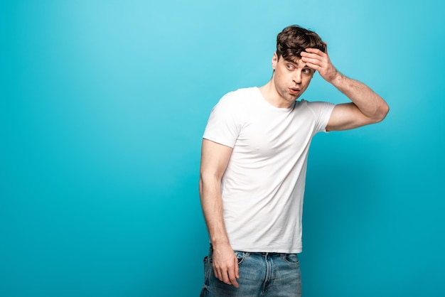 Jeune homme choqué regardant loin tout en tenant la main sur la tête sur fond bleu