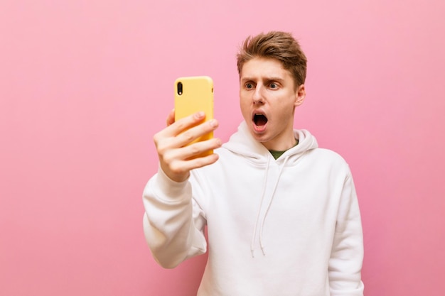 Un jeune homme choqué en sweat-shirt blanc se tient avec un smartphone à la main sur fond rose