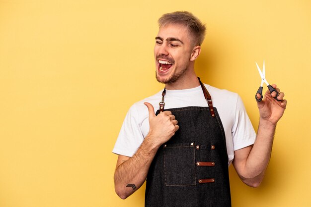 Jeune homme de coiffeur caucasien tenant des ciseaux isolés sur fond jaune pointe avec le pouce loin, riant et insouciant.