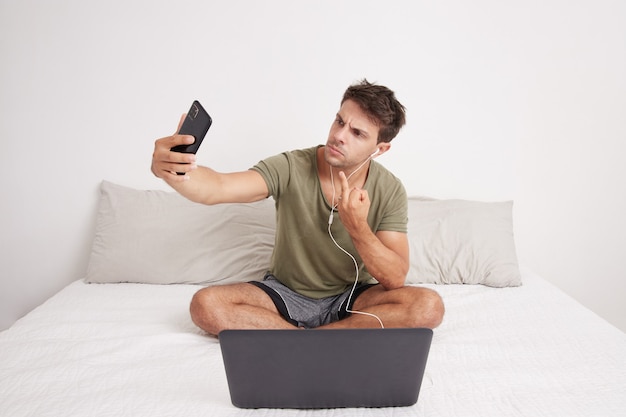 Jeune homme en colère prenant un selfie avec son téléphone portable assis sur son lit insultant avec son doigt. Il a son ordinateur portable et il le fait. Lit et chambre blanche.