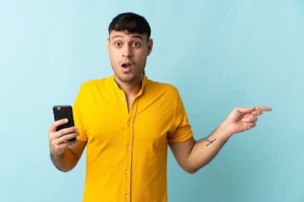 Jeune homme colombien à l'aide de téléphone mobile isolé sur bleu côté surpris et pointant