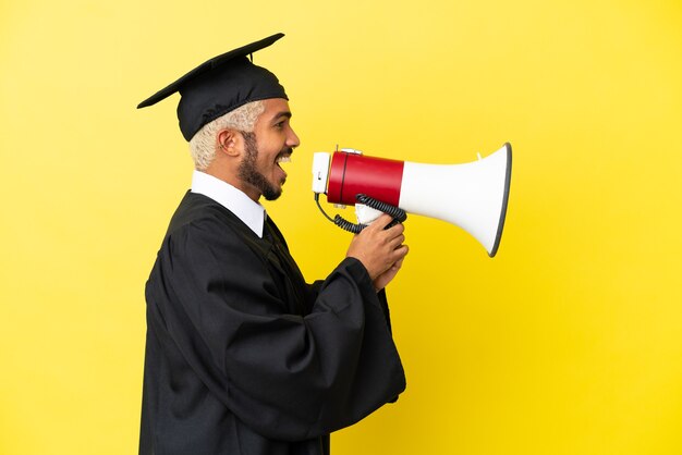 Jeune homme colombien diplômé universitaire isolé sur fond jaune criant à travers un mégaphone