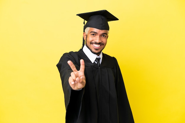 Jeune homme colombien diplômé universitaire isolé sur fond jaune souriant et montrant le signe de la victoire