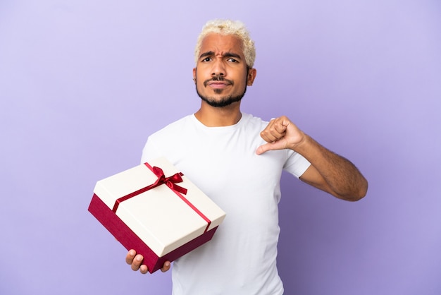 Jeune homme colombien tenant un cadeau isolé sur fond violet fier et satisfait de lui-même