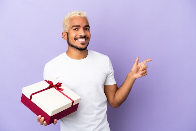 Jeune homme colombien tenant un cadeau isolé sur fond violet, pointant le doigt sur le côté