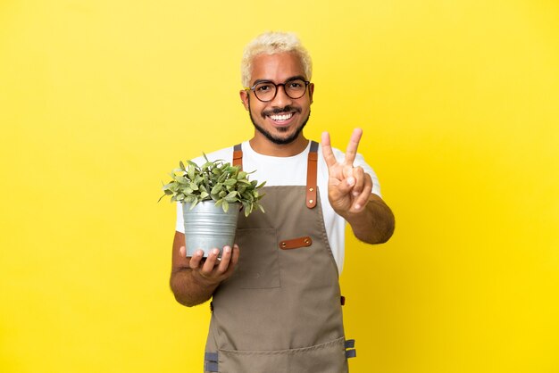 Jeune homme colombien tenant une plante isolée sur fond jaune souriant et montrant le signe de la victoire