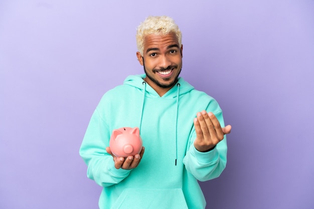 Jeune homme colombien tenant une tirelire isolée sur fond violet invitant à venir avec la main. Heureux que tu sois venu