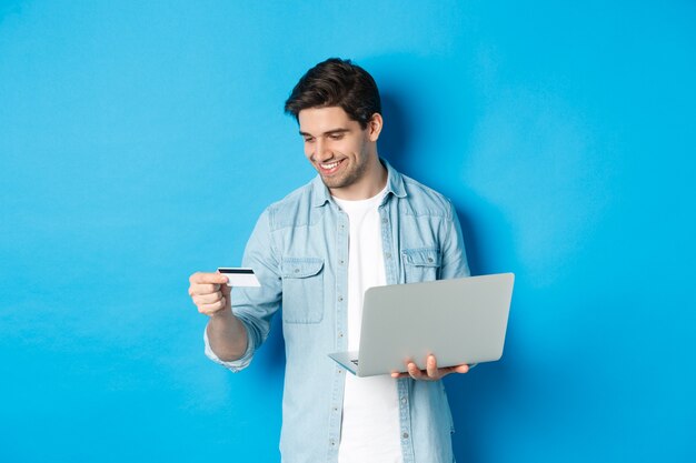 Jeune homme commande en ligne, tenant une carte de crédit et un ordinateur portable, faisant des achats sur Internet, debout sur fond bleu