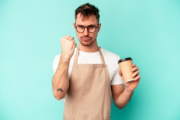 Jeune homme de commis de magasin tenant un café isolé sur fond bleu montrant le poing à la caméra, expression faciale agressive.