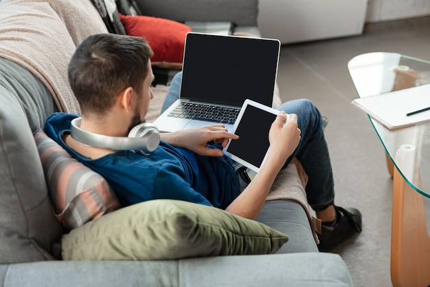 Jeune homme concentré étudiant à la maison pendant des cours en ligne ou des informations gratuites par lui-même. Devient développeur de logiciels, responsable informatique tout en étant isolé, mis en quarantaine. Utilisation d'un ordinateur portable, d'un smartphone, d'un casque.