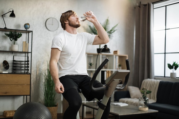 Jeune homme concentré portant des vêtements de sport buvant de l'eau à l'aide d'un vélo d'exercice