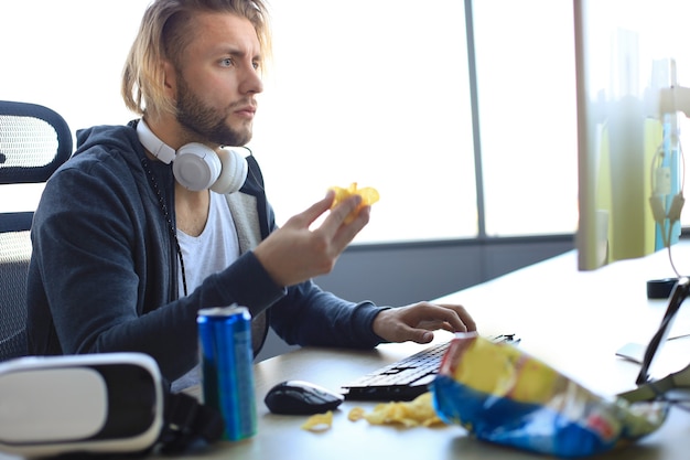 Jeune homme concentré vêtu de vêtements décontractés utilisant un ordinateur, diffusant une vidéo en streaming ou une vidéo et mangeant des chips.