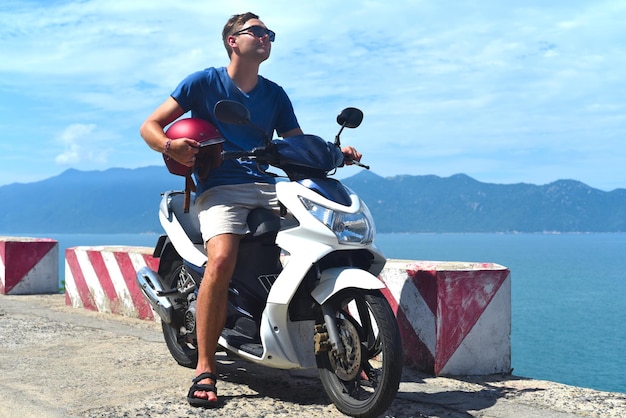 Jeune homme conducteur de moto assis et tenant un casque contre la mer de Chine du Sud au Vietnam