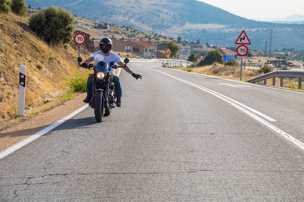 Jeune homme conduisant une moto sur la route en montagne en journée ensoleillée tout en faisant les salutations de motards.