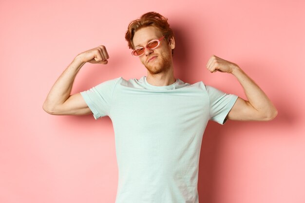 Jeune homme confiant aux cheveux rouges portant des lunettes de soleil d'été et un t-shirt montrant un corps fort et en forme...