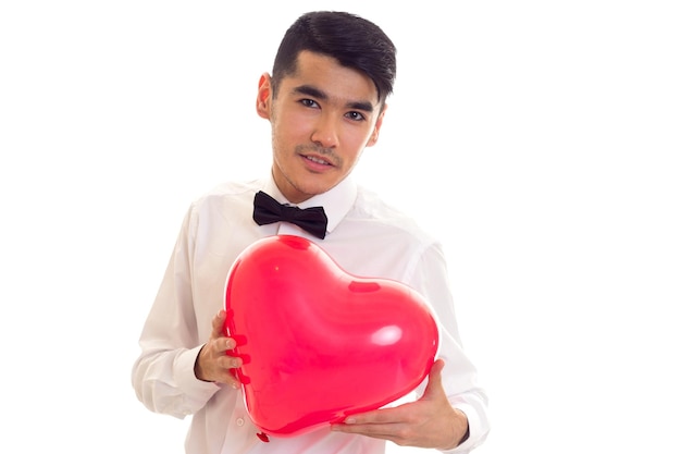 Jeune homme confiant en chemise blanche avec noeud papillon noir tenant un ballon en forme de coeur rouge sur fond blanc en studio