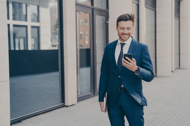 Jeune homme confiant et heureux consultant en affaires tenant un smartphone moderne et souriant à l'extérieur