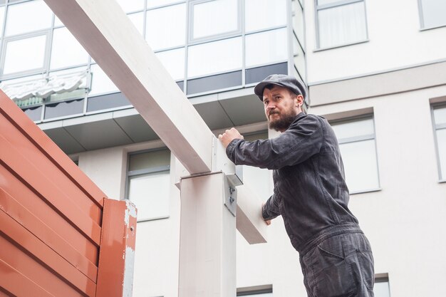 Un jeune homme avec un constructeur de barbe dans un uniforme noir de construction, un chapeau gris construit une maison d'une poutre en bois