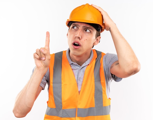 Jeune homme de constructeur impressionné en uniforme pointe vers le haut isolé sur mur blanc