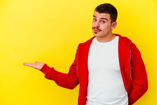 Jeune homme cool caucasien isolé sur un mur jaune montrant un espace de copie sur une paume et tenant une autre main sur la taille.