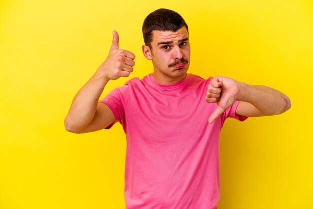 Jeune homme cool caucasien isolé sur un mur jaune montrant les pouces vers le haut et les pouces vers le bas, difficile de choisir le concept