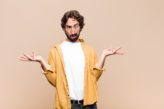 Jeune homme cool fou haussant les épaules avec une expression stupide, folle, confuse, perplexe, se sentant agacé et désemparé contre le mur plat